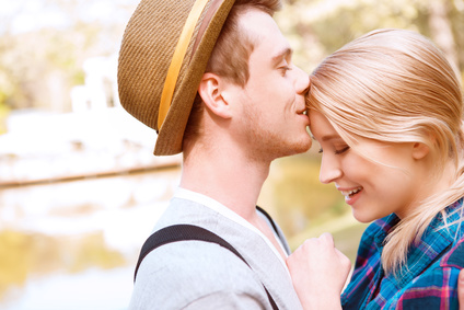 Sweet kiss. Handsome young man standing in park near lake and kissing his girlfriend on forehead