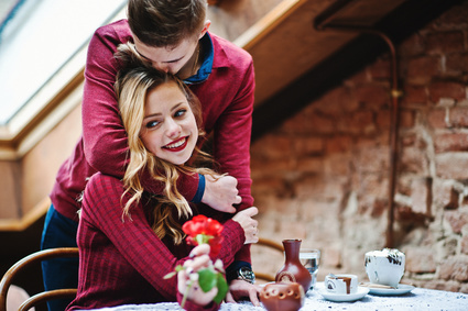 Young beautiful stylish couple in a red dress in love story at the vintage cafe with big windows at the roof, boy gives flower as present for girlfriend